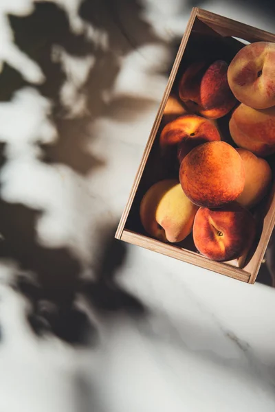 Top view of wooden box full of fresh peaches on light marble surface with shadows — Stock Photo