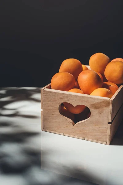 Composición de alimentos con albaricoques en caja de madera con signo en forma de corazón en superficie clara con fondo negro - foto de stock