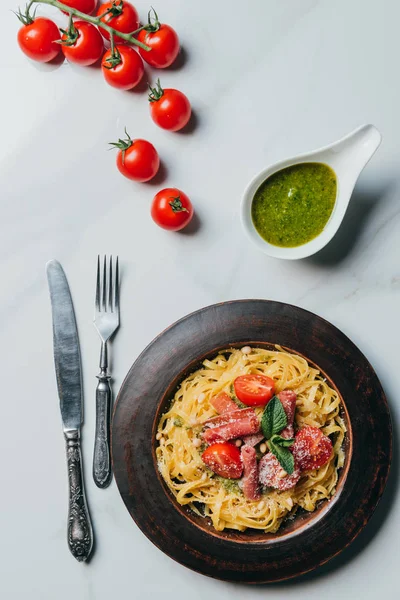 Vista dall'alto pasta con foglie di menta, marmellata e pomodorini ricoperti da parmigiano su piatto di marmo tavolo con pesto in ciotola, coltello e forchetta — Foto stock