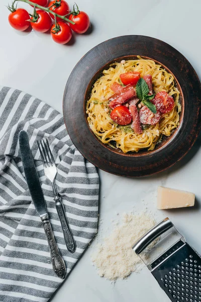Top view of pasta with mint leaves, jamon and cherry tomatoes covered by parmesan on plate at marble table with kitchen towel, knife, fork, cheese and grater — Stock Photo