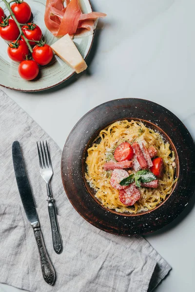 Vista elevada de platos con pasta y jamón, tomates parmesano y cereza sobre mesa de mármol con toalla de cocina, tenedor y cuchillo - foto de stock