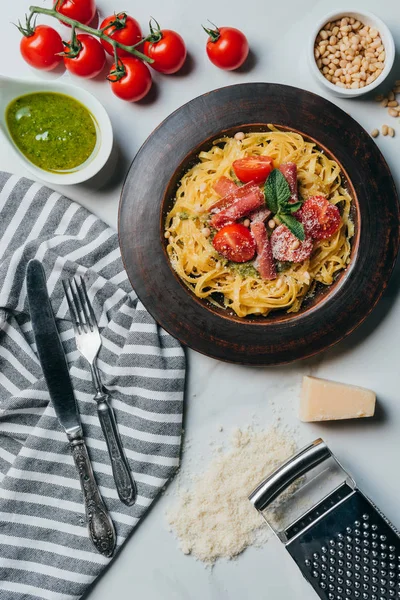 Pasta with mint leaves, jamon and cherry tomatoes covered by grated parmesan on plate at marble table with kitchen towel, knife, grater, cheese, fork, pine nuts and pesto sauce in bowl — Stock Photo