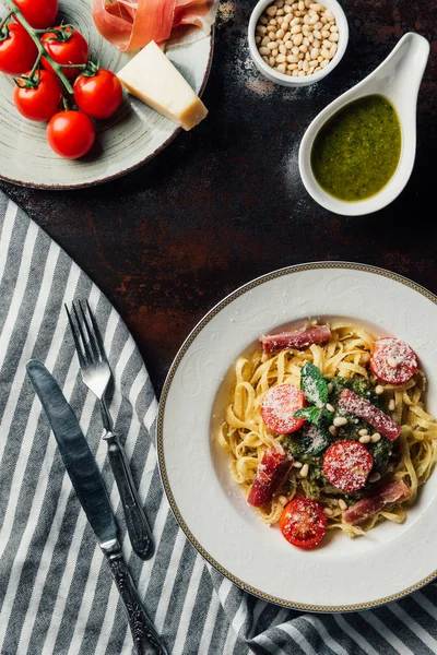 Top view of pasta with mint leaves, jamon and cherry tomatoes covered by grated parmesan on plate at table with kitchen towel, knife, fork, pine nuts and pesto sauce in bowl — Stock Photo