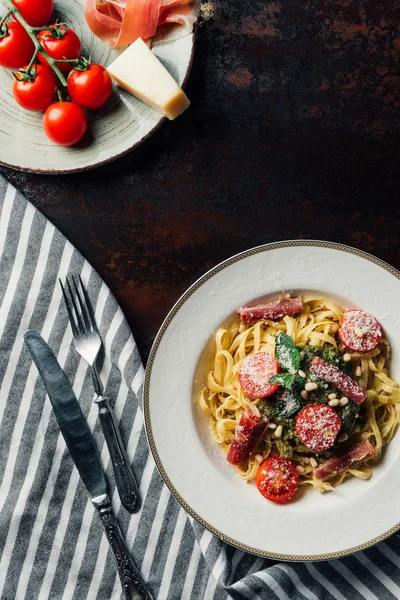 Vista de cima placas com macarrão e jamon, parmesão e jamon na mesa com toalha de cozinha, garfo e faca — Fotografia de Stock