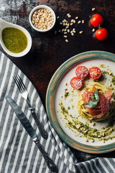 Top view of pasta with mint leaves, jamon and cherry tomatoes covered by parmesan on plate at table with kitchen towel, knife, fork, pine nuts and pesto in bowl — Stock Photo