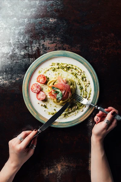 Imagen recortada de la mujer comiendo pasta con hojas de menta, jamón y tomates cherry cubiertos por parmesano rallado en la mesa - foto de stock