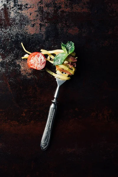 Elevated view of fork wrapped by pasta with mint leaves, jamon and cherry tomatoes covered by grated parmesan on table — Stock Photo