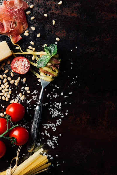 Vista superior de tenedor envuelto por pasta con hojas de menta, jamón, piñones y tomates cherry cubiertos de parmesano rallado en la mesa rodeado de ingredientes - foto de stock