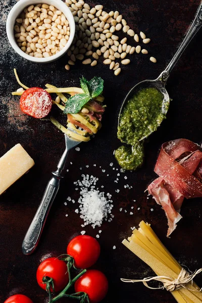 Elevated view of fork wrapped by pasta surrounded by pine nuts, pesto, parmesan, raw spaghetti, cherry tomatoes, salt and jamon on table — Stock Photo