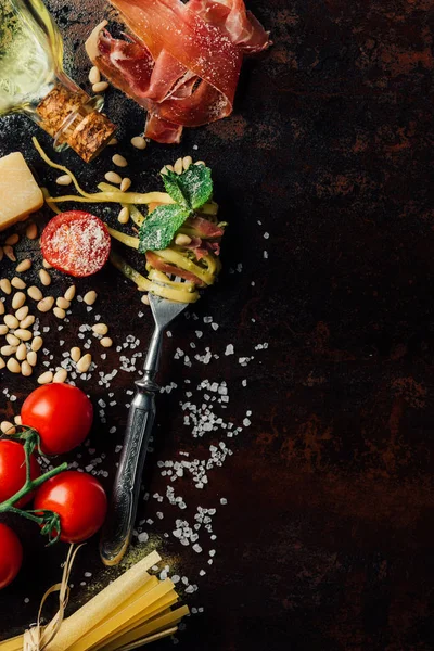 Vista desde arriba tenedor envuelto por pasta rodeada de piñones, pesto, parmesano, espaguetis crudos, tomates cherry, sal, aceite de oliva y jamón en la mesa — Stock Photo