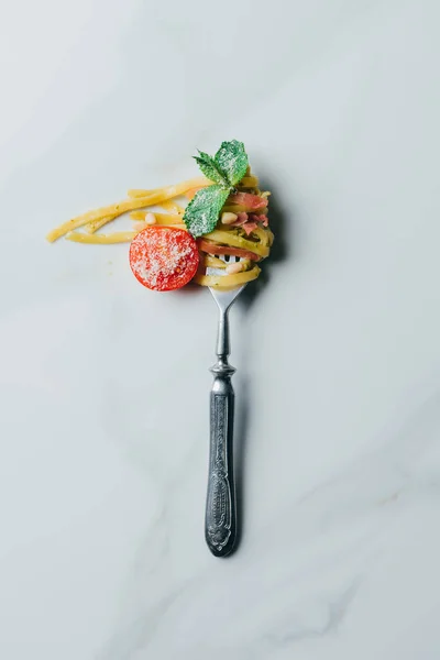 Top view of fork wrapped by pasta with mint leaves, jamon and cherry tomatoes covered by grated parmesan on marble table — Stock Photo
