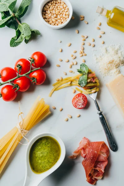 Elevated view of fork wrapped by pasta surrounded by pine nuts, pesto, parmesan, raw spaghetti, cherry tomatoes, salt, olive oil and jamon on marble table — Stock Photo