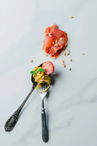 Vue surélevée sur cuillère et fourchette enveloppée de pâtes aux feuilles de menthe, pignons de pin, confiture et tomates cerises recouvertes de parmesan râpé sur table en marbre — Photo de stock