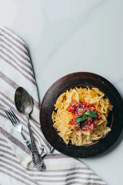 Vista desde arriba de pasta con hojas de menta y salsa cubierta de parmesano rallado sobre mesa de mármol con tenedor, cuchillo y toalla de cocina - foto de stock