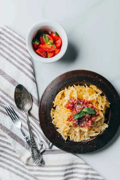 Повышенный вид тарелки с макаронами, кухонным полотенцем, вилкой, ложкой и миской с помидорами черри на мраморном столе — стоковое фото