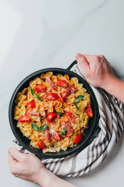 Cropped image of man holding pan with pasta covered by grated parmesan with cherry tomatoes, jamon, mint leaves over marble table — Stock Photo