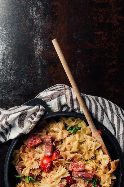 Elevated view of pasta with jamon, cherry tomatoes, mint leaves covered by grated parmesan in pan with wooden spatula on table with kitchen towel — Stock Photo