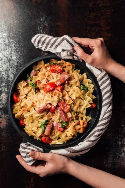 Cropped image of woman holding pasta with jamon, cherry tomatoes, mint leaves covered by grated parmesan in pan wrapped by kitchen towel over table — Stock Photo