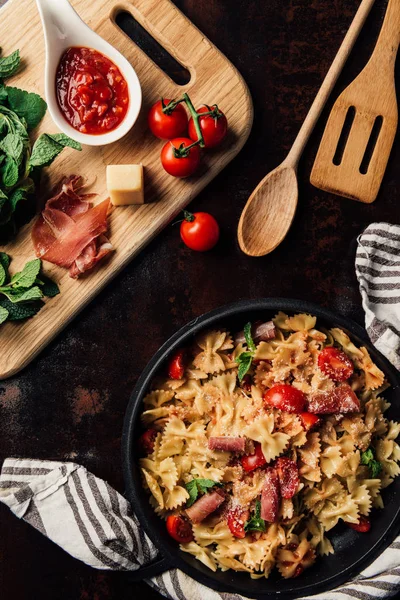 Elevated view of pasta with jamon, pine nuts, sauce, cherry tomatoes, mint leaves covered by grated parmesan in pan surrounded by kitchen towel and cutting board with ingredients on table — Stock Photo