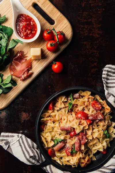 Top view of pasta with jamon, pine nuts, sauce, cherry tomatoes, mint leaves covered by grated parmesan in pan surrounded by kitchen towel and cutting board with ingredients on table — Stock Photo