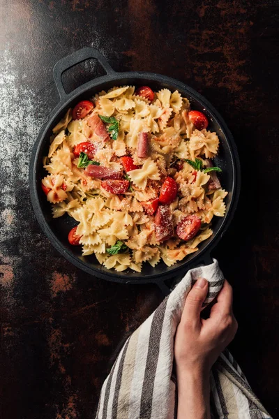 Partial view of woman holding pasta with jamon, cherry tomatoes, mint leaves covered by grated parmesan in pan wrapped by kitchen towel over table — Stock Photo