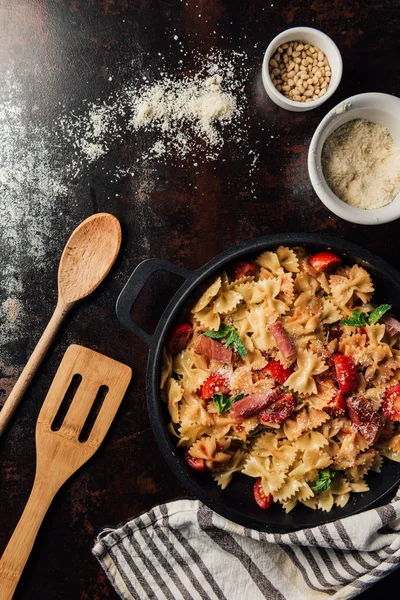 View from above of pasta with jamon, pine nuts, sauce, cherry tomatoes, mint leaves covered by grated parmesan in pan surrounded by kitchen towel, wooden spatula and spoon on table — Stock Photo