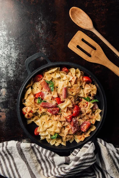 Elevated view of pasta with jamon, cherry tomatoes, mint leaves covered by grated parmesan in pan on table with kitchen towel, wooden spoon and spatula — Stock Photo
