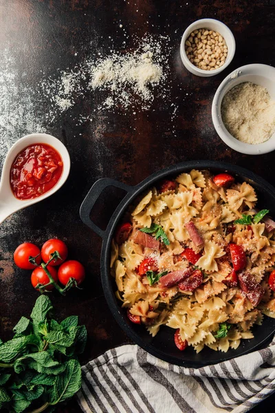 Vista de cima de massas com jamon, pinhões, molho, tomate cereja, folhas de hortelã cobertas por parmesão ralado na panela cercada por ingredientes e toalha de cozinha na mesa — Fotografia de Stock