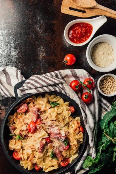 Top view of pasta with jamon, pine nuts, sauce, cherry tomatoes, mint leaves covered by grated parmesan in pan surrounded by ingredients and kitchen towel on table — Stock Photo