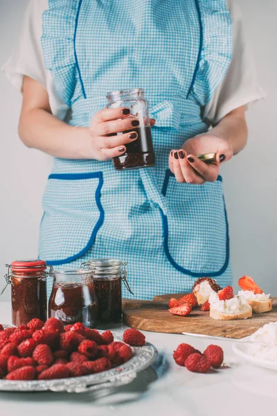 Imagen recortada de mujer en delantal sosteniendo frasco abierto con mermelada de frutas en la cocina - foto de stock