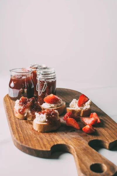 Vista ravvicinata del tagliere con marmellata di frutta in vasetti e panini con crema di formaggio e marmellata su grigio — Foto stock