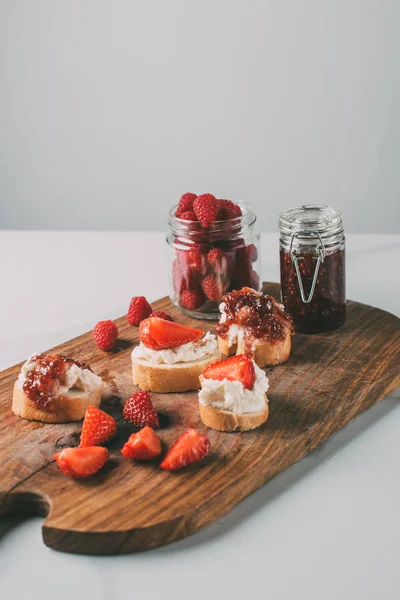 Nahaufnahme Schneidebrett mit Erdbeermarmelade in Gläsern und Sandwiches mit Frischkäse und Marmelade auf grau — Stockfoto