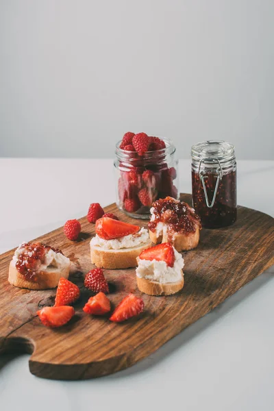 Close up tiro de tábua de corte com geléia de morango em frascos e sanduíches com queijo creme e geléia em cinza — Fotografia de Stock