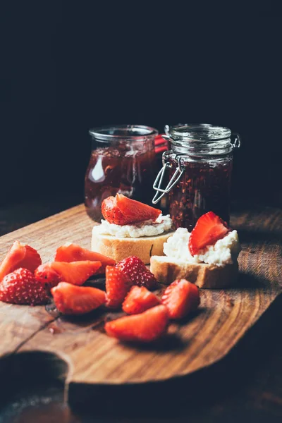 Selective focus of sandwiches with cream cheese, strawberry slices and fruit jam on cutting board on black — Stock Photo