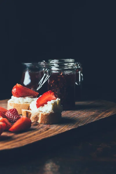 Selective focus of sandwiches with cream cheese, strawberry slices and fruit jam on cutting board on black — Stock Photo