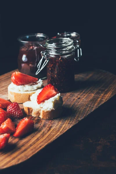 Selective focus of fruit jam in different jars, sandwiches with cream cheese, strawberry slices and fruit jam on cutting board on black — Stock Photo