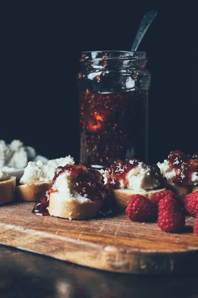 Vista de perto de framboesas, sanduíches com engarrafamento de fruto e queijo de creme em tábua de corte em preto — Fotografia de Stock