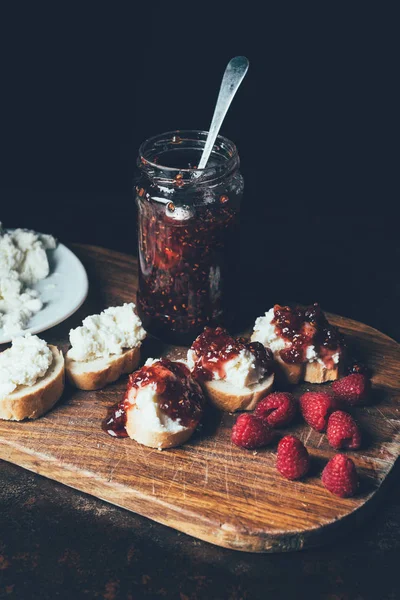 Vista elevada de frambuesas, sándwiches con mermelada de frutas y queso crema en la tabla de cortar en negro - foto de stock