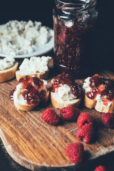 Close up view of raspberries, sandwiches with fruit jam and cream cheese on cutting board on black — Stock Photo