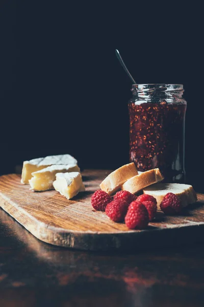 Nahaufnahme von Brie, Himbeeren, Marmelade im Glas und Baguette auf Schneidebrett am Tisch — Stockfoto