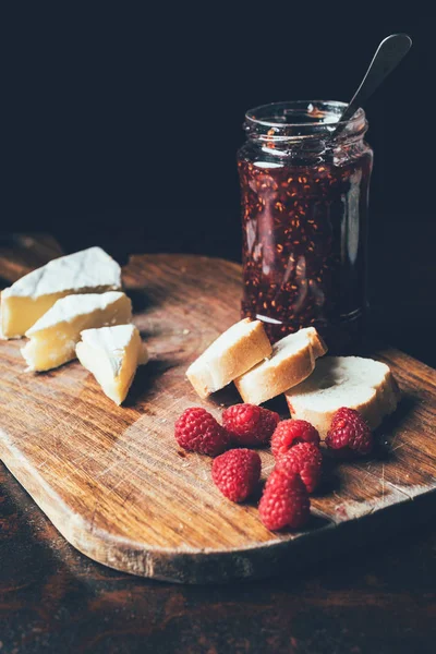 Selective focus of brie, raspberries, jam in jar and baguette on cutting board at table — Stock Photo