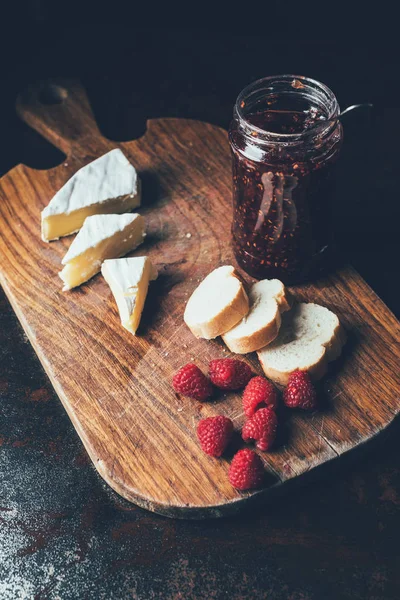 Erhöhte Ansicht von Brie, Himbeeren, Marmelade im Glas und Baguette auf Schneidebrett am Tisch — Stockfoto