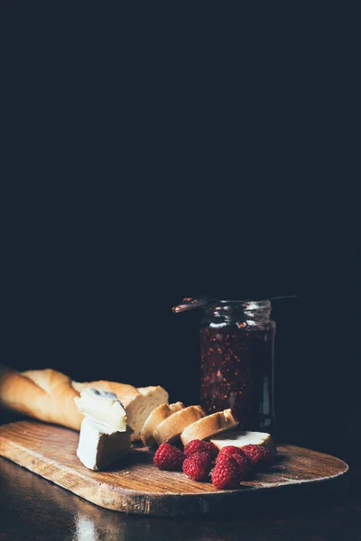 Vue de face de brie, framboises, confiture en pot et baguette sur planche à découper sur noir — Photo de stock