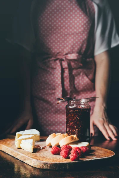 Imagen recortada de la mujer en delantal de pie cerca de la mesa con brie, rebanadas de baguette, frambuesas y tarro de mermelada en la tabla de cortar - foto de stock