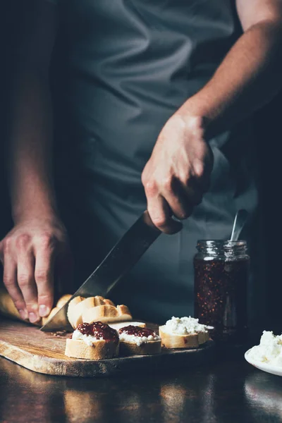 Imagen recortada de hombre en delantal corte baguette para hacer sándwiches con queso crema y mermelada de frutas - foto de stock