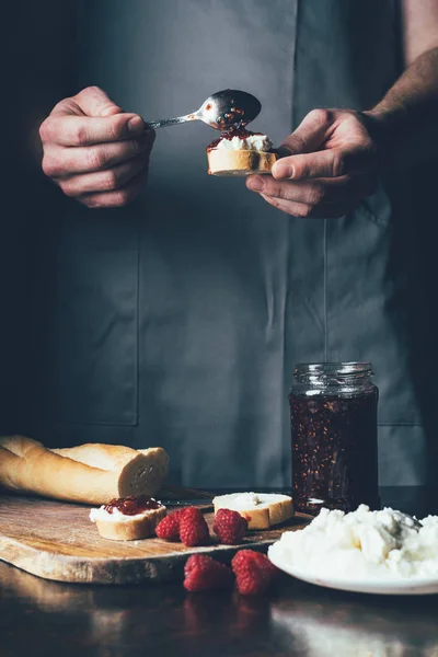 Vista parcial del hombre en delantal extendiendo mermelada de fresa en la baguette con queso crema — Stock Photo