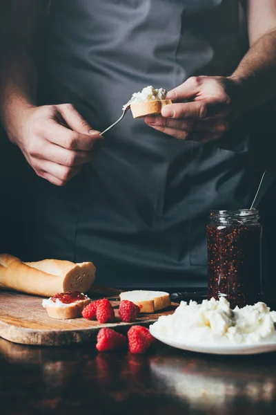 Imagen recortada del hombre en delantal que extiende el queso crema en la baguette sobre la mesa con mermelada de frutas — Stock Photo