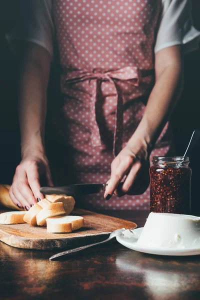 Abgeschnittenes Bild einer Frau in Schürze, die Baguette an Bord schneidet, in der Nähe von Frischkäse und Marmeladenglas am Tisch — Stockfoto