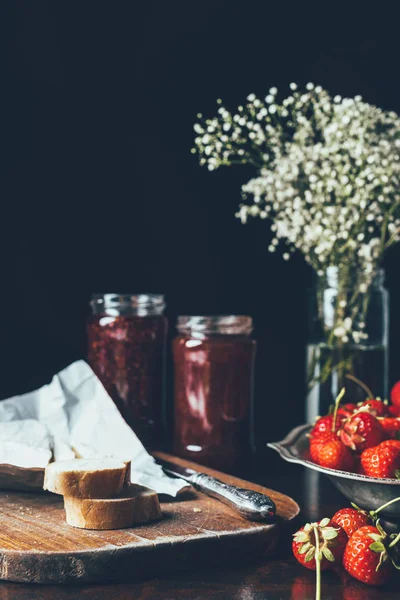 Close up view of strawberries with jam in jars and flowers on black — Stock Photo