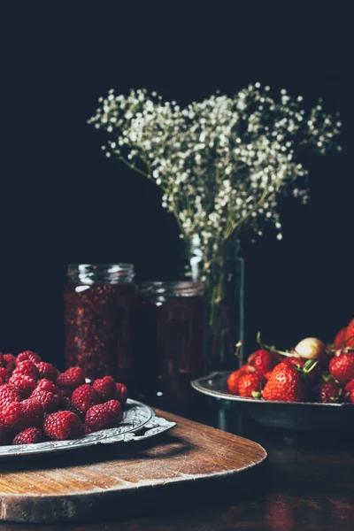 Selective focus of raspberries, strawberries, jars with jam and flowers on black — Stock Photo